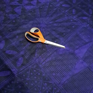 May include: An orange-handled pair of scissors rests on a patterned fabric. The fabric features a dark blue and purple geometric design with lines and shapes. The scissors are open, with silver blades.