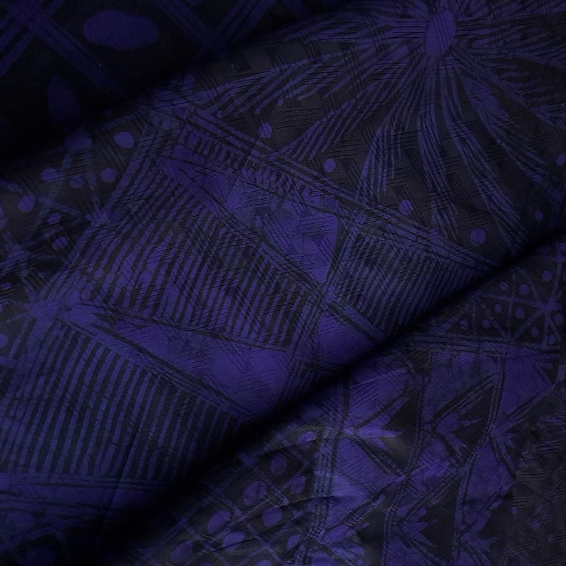 May include: Close-up of a folded fabric with a dark purple and black geometric pattern. The design includes lines, dots, and shapes, creating a textured appearance. The fabric has a slight sheen, suggesting a smooth material.