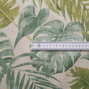 May include: Close-up of a beige fabric with a green tropical leaf print. A ruler is placed on the fabric to show the scale. The ruler is marked in inches.
