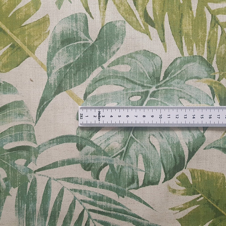 May include: Close-up of a beige fabric with a green tropical leaf print. A ruler is placed on the fabric to show the scale. The ruler is marked in inches.