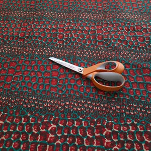 May include: Red and teal fabric with a repeating pattern of small, rounded shapes. A pair of orange-handled scissors is lying on top of the fabric.