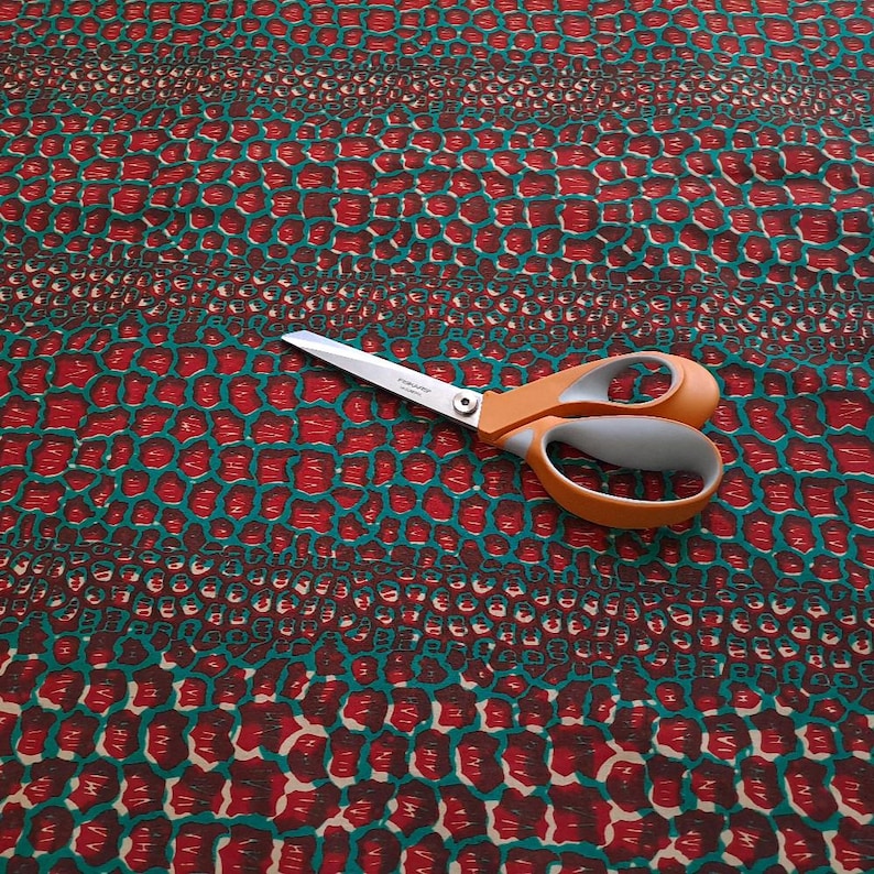 May include: Red and teal fabric with a repeating pattern of small, rounded shapes. A pair of orange-handled scissors is lying on top of the fabric.