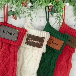 May include: Four personalized Christmas stockings with names on leather tags. The stockings are red, white, green, and red. The names on the tags are Henry, Alexander, Sophia, and Emma.