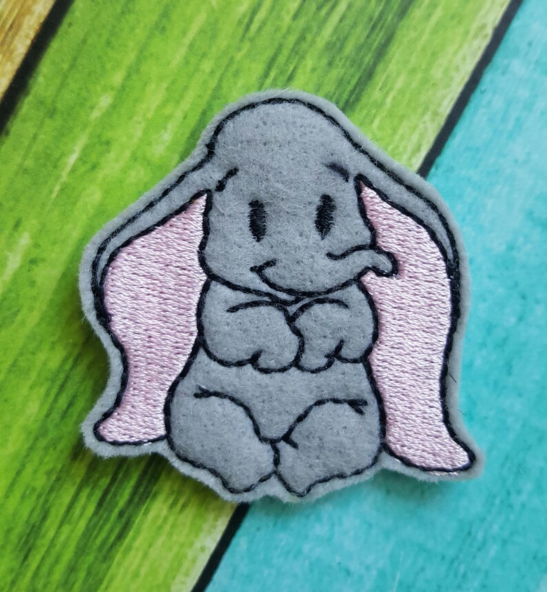 May include: A gray felt embroidered patch of Dumbo the elephant from Disney. Dumbo has large pink ears and is sitting with his trunk curled up.