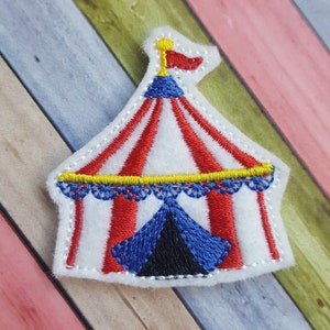 May include: A white felt applique of a red and white striped circus tent with a blue and yellow top and a red flag. The tent has a blue and black flap.