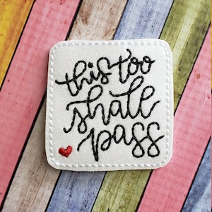 May include: White square patch with the embroidered phrase "this too shall pass" in black script. A small red heart is stitched below the text. The patch is on a colorful wooden background.