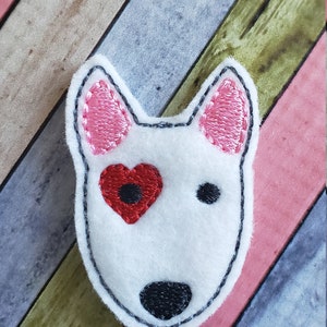May include: White felt dog applique with pink ears and a red heart for an eye. The applique is on a striped wooden background.