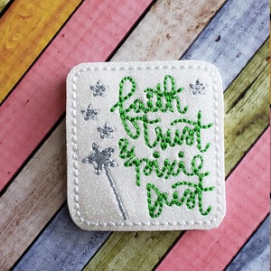 May include: White fabric square with green embroidered text that reads "Faith Trust & Pixie Dust" with silver glitter stars and a silver glitter wand.