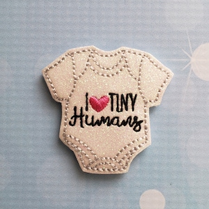May include: White glitter iron-on patch shaped like a baby onesie with the text "I love tiny humans" and a red heart.
