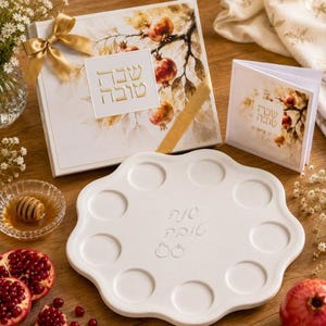 May include: A white ceramic serving tray with eight circular indentations and Hebrew text, accompanied by a gift box and a folded card featuring pomegranate illustrations and gold ribbon accents, alongside a small glass bowl of honey.