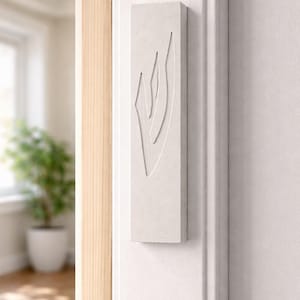 May include: A rectangular, off-white mezuzah case with a carved design, mounted on a white wall. The design features a stylized, abstract shape. The case is positioned near a wooden door frame.