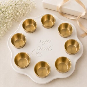 May include: A white, scalloped-edge serving tray featuring eight circular, brushed-gold metal bowls arranged in a ring. The center of the tray is embossed with Hebrew text and two small pomegranate icons, creating a decorative display piece.