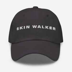 May include: A dark gray baseball cap with the embroidered text "SKIN WALKER" in white. The cap has a curved brim and a button on top.