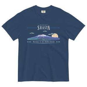 May include: Navy blue t-shirt featuring a graphic of Mount Shasta with UFOs, a full moon, and the text "Mount Shasta" and "Doorway to the Hidden Realm". The design is in shades of blue, white, and yellow.