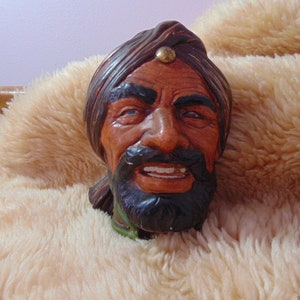 May include: A ceramic figurine of a smiling man's head with a turban. The face is painted with brown, red, and black tones, with blue eyes and a gold accent on the turban. The figurine rests on a fluffy, light-colored surface.