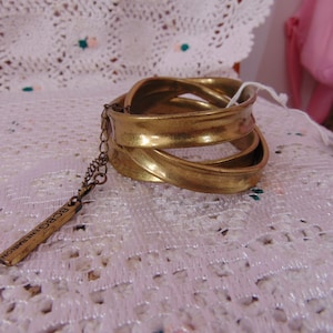 May include: A gold-toned metal cuff bracelet with a layered design. The bracelet has a textured surface and a dangling tag with the inscription "BCBG Generation". The bracelet is displayed on a white lace surface.