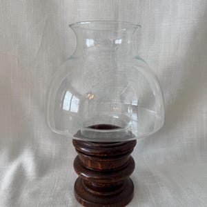 K&ouml;nnte beinhalten: Ein klarer Glas-Windlichtschirm sitzt auf einem dunkelbraunen Holzsockel. Der Sockel besteht aus gestapelten, kreisf&ouml;rmigen Holzst&uuml;cken. Der Glasschirm hat eine abgerundete Oberseite und eine ausgestellte &Ouml;ffnung.
