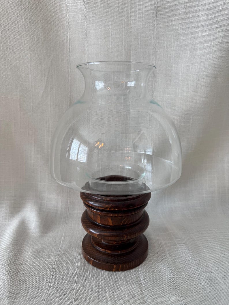 K&ouml;nnte beinhalten: Ein klarer Glas-Windlichtschirm sitzt auf einem dunkelbraunen Holzsockel. Der Sockel besteht aus gestapelten, kreisf&ouml;rmigen Holzst&uuml;cken. Der Glasschirm hat eine abgerundete Oberseite und eine ausgestellte &Ouml;ffnung.