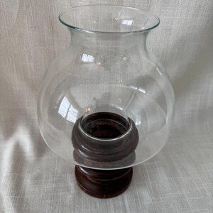 K&ouml;nnte beinhalten: Ein klarer Glas-Windlicht-Kerzenhalter mit einer runden, bauchigen Form. Das Glas sitzt auf einem dunkelbraunen, geschnitzten Holzsockel. Der Sockel hat ein mehrstufiges Design. Der Kerzenhalter ist so konzipiert, dass er eine Kerze vor Zugluft sch&uuml;tzt.