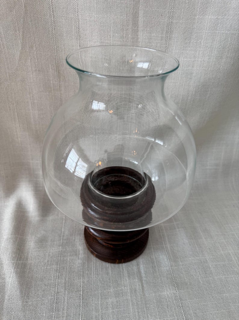 K&ouml;nnte beinhalten: Ein klarer Glas-Windlicht-Kerzenhalter mit einer runden, bauchigen Form. Das Glas sitzt auf einem dunkelbraunen, geschnitzten Holzsockel. Der Sockel hat ein mehrstufiges Design. Der Kerzenhalter ist so konzipiert, dass er eine Kerze vor Zugluft sch&uuml;tzt.