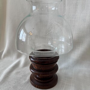 K&ouml;nnte beinhalten: Ein klarer Glas-Windlichtschirm sitzt auf einem dunkelbraunen Holzsockel. Der Sockel besteht aus gestapelten, abgerundeten Holzst&uuml;cken. Der Glasschirm hat eine abgerundete Oberseite und eine ausgestellte &Ouml;ffnung.