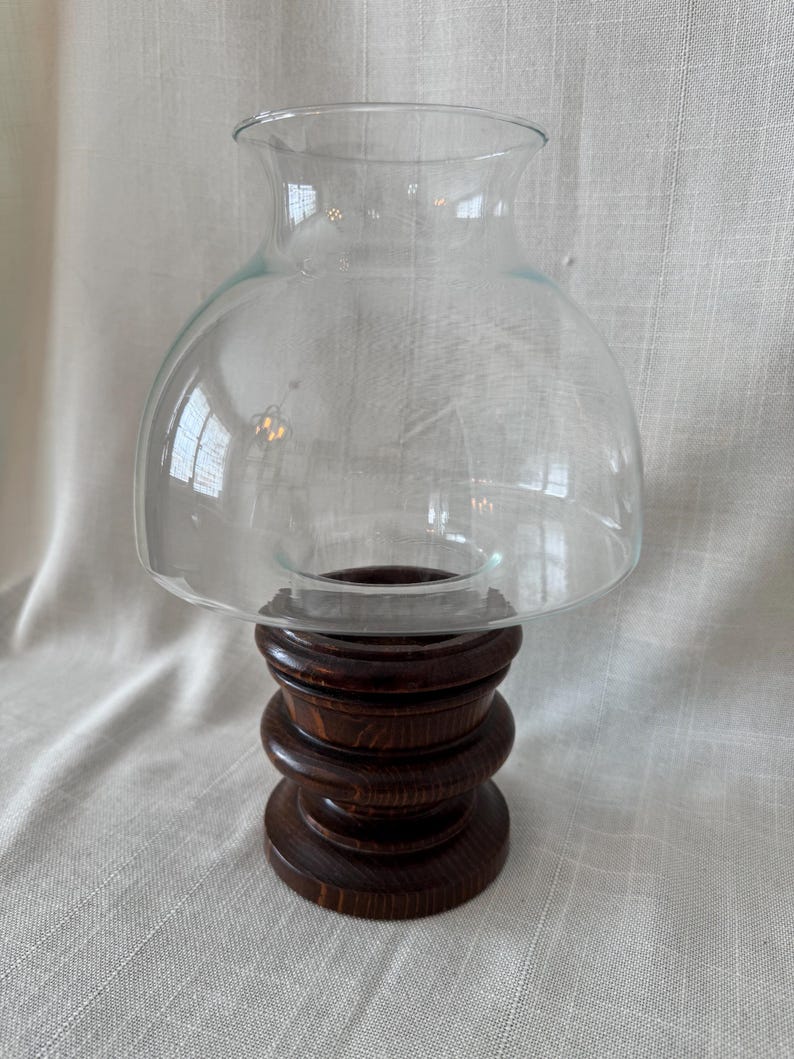 K&ouml;nnte beinhalten: Ein klarer Glas-Windlichtschirm sitzt auf einem dunkelbraunen Holzsockel. Der Sockel besteht aus gestapelten, abgerundeten Holzst&uuml;cken. Der Glasschirm hat eine abgerundete Oberseite und eine ausgestellte &Ouml;ffnung.
