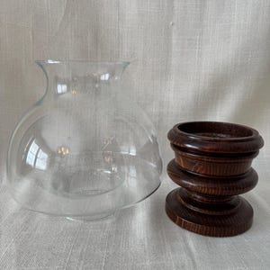 K&ouml;nnte beinhalten: Eine transparente Glasglocke mit rundem K&ouml;rper und ausgestellter &Ouml;ffnung, neben einem dunkelbraunen Holzsockel. Der Sockel hat ein mehrschichtiges Design mit kreisf&ouml;rmiger Ober- und Unterseite. Die Glocke und der Sockel dienen wahrscheinlich dekorativen Zwecken.