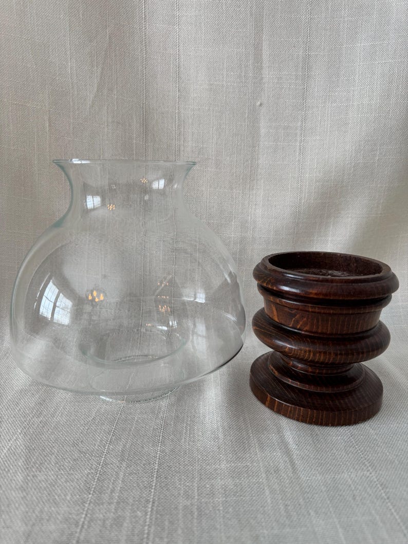 K&ouml;nnte beinhalten: Eine transparente Glasglocke mit rundem K&ouml;rper und ausgestellter &Ouml;ffnung, neben einem dunkelbraunen Holzsockel. Der Sockel hat ein mehrschichtiges Design mit kreisf&ouml;rmiger Ober- und Unterseite. Die Glocke und der Sockel dienen wahrscheinlich dekorativen Zwecken.