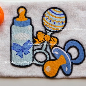 Peut inclure: Une serviette blanche avec des articles de bébé brodés, dont un biberon bleu, un hochet bleu et jaune et une sucette bleue. La serviette a également un hochet jaune et orange.