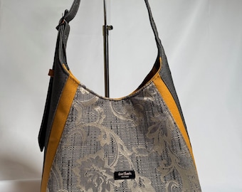 May include: A shoulder bag with a floral pattern in gray and beige. The bag has a black strap and trim with a yellow accent. The bag has the text "Give Thanks" on a small black label.