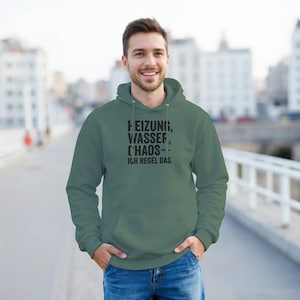 May include: Sage green hoodie with the text "HEIZUNG, WASSER, CHAOS - ICH REGEL DAS." in black. The hoodie has a drawstring hood and a front pocket. The person is wearing the hoodie.