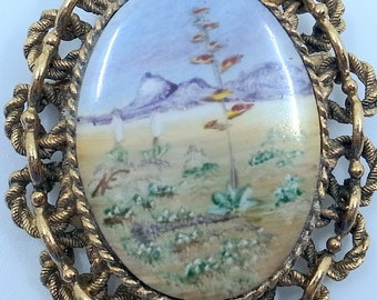 May include: A gold-toned pendant necklace with a delicate, ornate frame. The pendant features a hand-painted porcelain oval with a desert landscape scene. The scene includes a tall, flowering plant, mountains, and other desert flora.