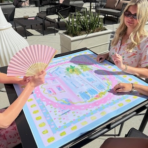 May include: A colorful, square decorative mat with a pastel illustration of a building, surrounded by a pink border and a light blue background. A pink hand fan is visible. The mat is on a black table.