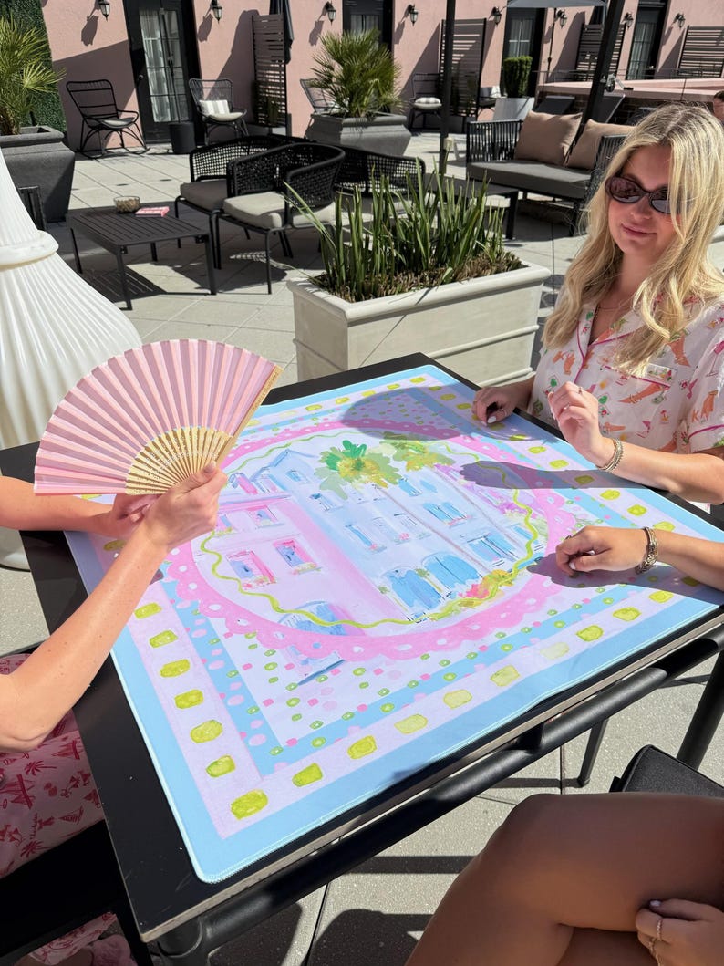 May include: A colorful, square decorative mat with a pastel illustration of a building, surrounded by a pink border and a light blue background. A pink hand fan is visible. The mat is on a black table.