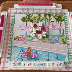 May include: A Mahjong set with a tropical pool scene design. The game board features a pink and white checkered border and a painted illustration of a pool with lounge chairs, umbrellas, and palm trees. The set includes pink tiles and racks.