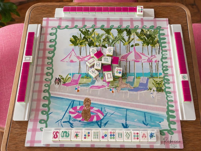 May include: A Mahjong set with a tropical pool scene design. The game board features a pink and white checkered border and a painted illustration of a pool with lounge chairs, umbrellas, and palm trees. The set includes pink tiles and racks.