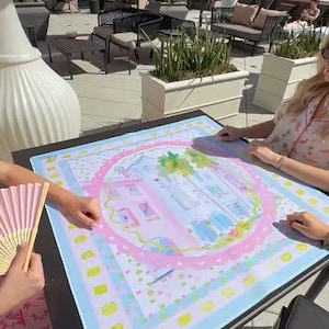 May include: A colorful, square art print featuring a watercolor illustration of a building with palm trees and a pink border. The print is on a black table, with a pink and yellow fan in the foreground.