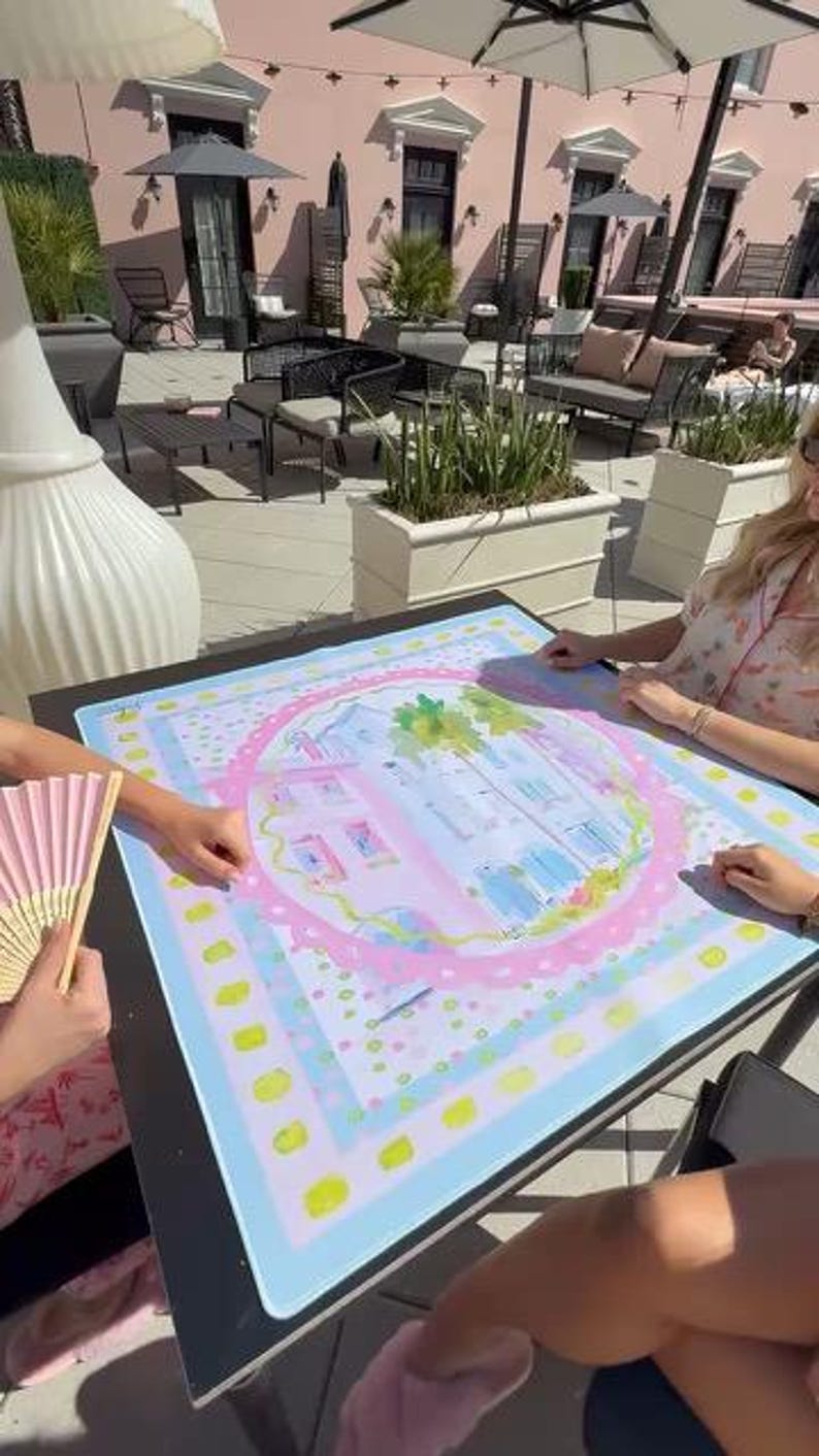 May include: A colorful, square art print featuring a watercolor illustration of a building with palm trees and a pink border. The print is on a black table, with a pink and yellow fan in the foreground.