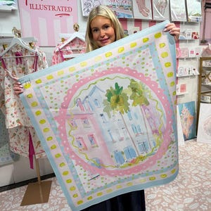May include: A square scarf with a watercolor illustration of buildings and palm trees, framed by a pink and blue border. The scarf has a light blue background with yellow accents. The scarf is held up in front of a display with the text "Rebecca Illustrated".