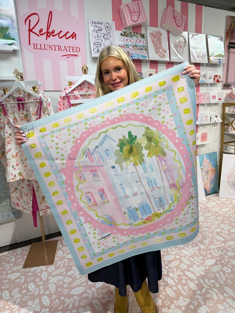 May include: A square scarf with a watercolor illustration of buildings and palm trees, framed by a pink and blue border. The scarf has a light blue background with yellow accents. The scarf is held up in front of a display with the text "Rebecca Illustrated".