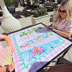 May include: A colorful, rectangular table mat with a pool scene design. The mat features a pink and white checkered border, a blue pool, and illustrations of people in a pool. The mat has the text "Rebecca & Co" in the corner.