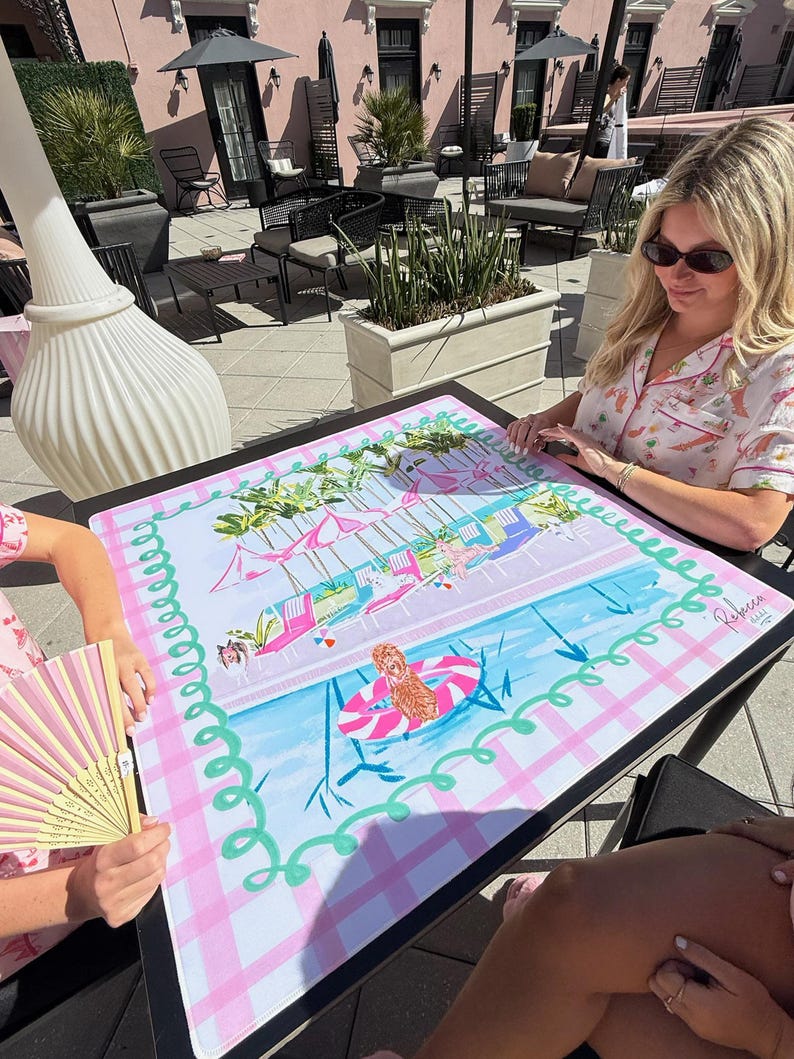 May include: A colorful, rectangular table mat with a pool scene design. The mat features a pink and white checkered border, a blue pool, and illustrations of people in a pool. The mat has the text "Rebecca & Co" in the corner.