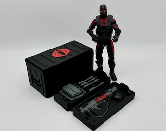 May include: A black plastic storage box with a red Cobra logo on the lid. The box is open and contains a variety of black plastic weapons and accessories. A black and red action figure is standing next to the box.