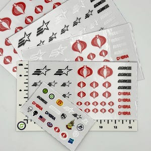 May include: Multiple sheets of G.I. Joe and Cobra themed adhesive decals featuring red Cobra logos, black star graphics, and text reading G.I. JOE and COBRA. A white ruler with black markings is visible, showing a width of approximately 14 inches.