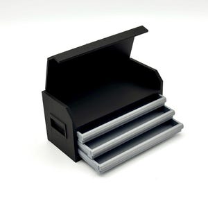 May include: A black storage box with three gray drawers. The top of the box is open, revealing a black interior. The drawers are pulled out, showcasing their contents. The box has a handle on the side.