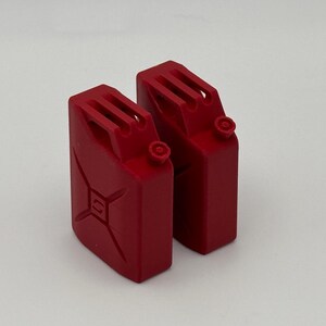 Op de afbeelding: Twee miniatuur rode brandstofkanisters met een klassiek jerrycan-ontwerp, voorzien van een handvat aan de bovenzijde, een kleine ronde dop voor de tuit en een verhoogd X-vormig structureel detail op de zijpanelen in mat rood.