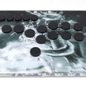 Może przedstawiać: Czarno-biały panel sterowania do gier arcade z wirującym wzorem. Panel ma 17 czarnych przycisków i napis "ERAEDUS" w prawym dolnym rogu.