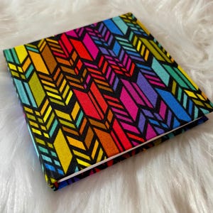 May include: A square fabric-covered notebook featuring a vibrant, repeating geometric arrow pattern in bright shades of red, orange, yellow, green, blue, and purple, all set against a stark black background with clean, sharp edges.
