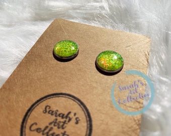 Op de afbeelding: Een paar groene glitter oorbellen met zilveren beslag. De oorbellen worden getoond op een bruine kaart met de tekst "Sarah's Art Collective" erop gedrukt.