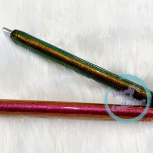 May include: Two glitter pens, one green and gold, the other pink and gold. Both pens have black tips.